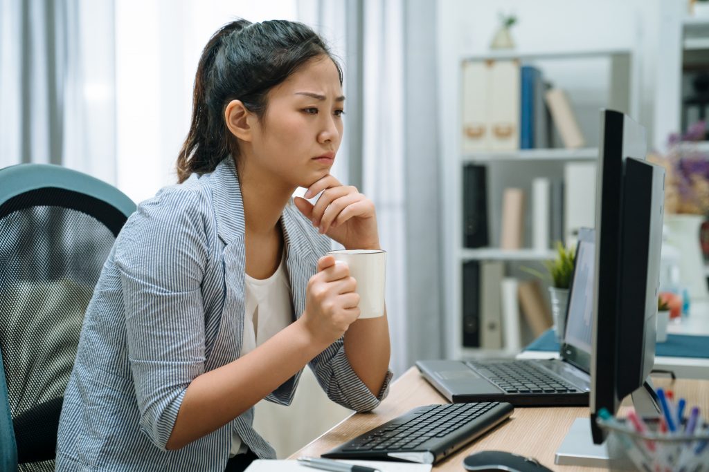 Pensive asian chinese woman in smart casual with desktop computer looking at screen holding coffee cup at home office. young thoughtful lady employee sitting at pc brainstorming during drink tea.